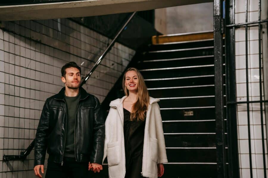 Two people stand on a subway stairwell, holding hands; a man in a black leather jacket and a woman in a white coat smile at the camera.